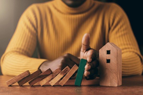 Pessoa interrompe a queda de peças de madeira tipo dominó com a mão, protegendo uma pequena casa de madeira ao lado.